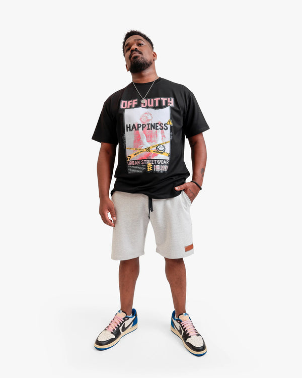 Front photo of an African American male model in a light grey backdrop wearing 'What Is Happiness' black relaxed t-shirt from Off Dutty's 'Rare and Radiant' collection, paired with Off Dutty White Short "Signature Brown Leather Logo Detail on Left" and Travis Scott Air Jordans. He accessorizes with necklace, bracelet, and showcases arm tattoos, posing with hand in pocket.