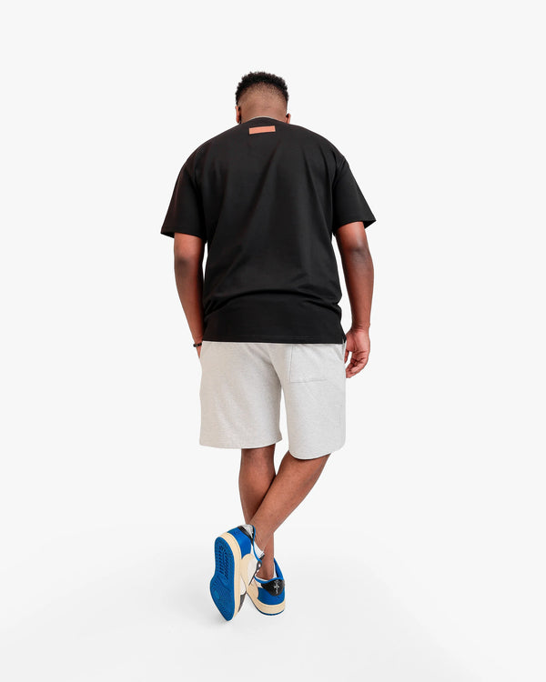 Back photo of an African American male model in a light grey backdrop wearing 'What Is Happiness' black relaxed t-shirt from Off Dutty's 'Rare and Radiant' collection, paired with Off Dutty White Short "Signature Brown Leather Logo Detail on Left" and Travis Scott Air Jordans. He accessorizes with necklace, bracelet, and posing with hand in pocket.