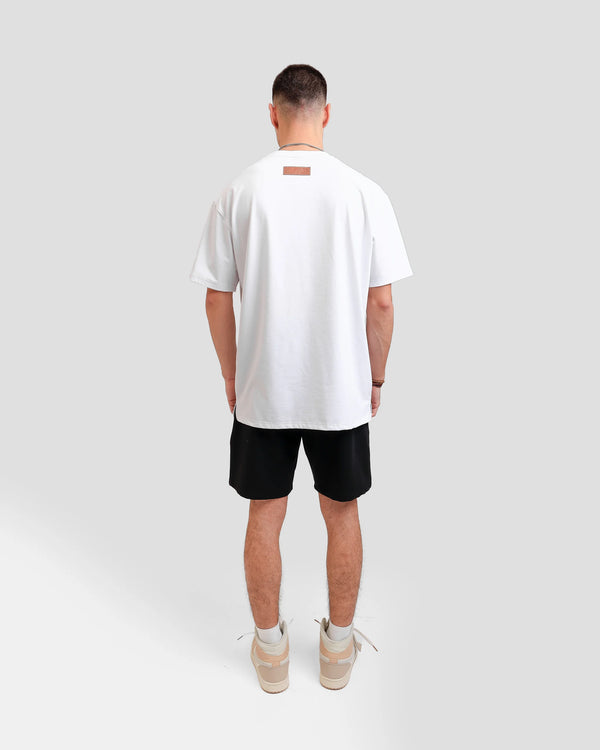 Back photo of a white Turkish look a like male model in a light grey backdrop wearing 'What Is Happiness' white relaxed T-shirt from Off Dutty's 'Rare and Radiant' collection, paired with Off Dutty Black Short "Signature Brown Leather Logo Detail on Left" and an Air Jordan 1 Mid 'Milan' sneaker. He accessorizes with necklaces, three bracelets on his right hand.