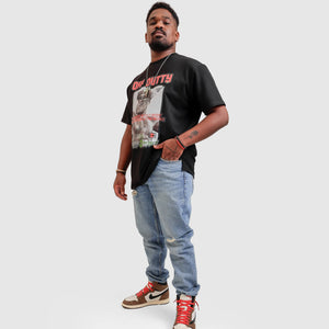 Front profile photo of an African American male model in a light grey backdrop wearing 'Embracing Your Presence' Black relaxed T-shirt from Off Dutty's 'Rare and Radiant' collection, paired with striped jeans and Travis Scott Air Jordans. He accessorizes with necklaces, red and black bracelets, and showcases arm tattoos, posing with hand in pocket.