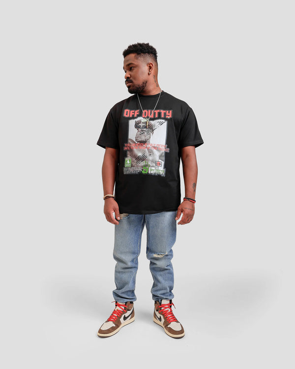 Front photo of an African American male model in a light grey backdrop wearing 'Embracing Your Presence' Black relaxed T-shirt from Off Dutty's 'Rare and Radiant' collection, paired with striped jeans and Travis Scott Air Jordans. He accessorizes with necklaces, red and black bracelets, and showcases arm tattoos, posing with head looking to the side.