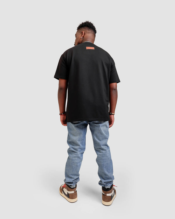 back photo of an African American male model in a light grey backdrop wearing 'Embracing Your Presence' Black relaxed T-shirt from Off Dutty's 'Rare and Radiant' collection, paired with striped jeans and Travis Scott Air Jordans. He accessorizes with necklaces, red and black bracelets, and showcases arm tattoos, posing with head looking to the right.