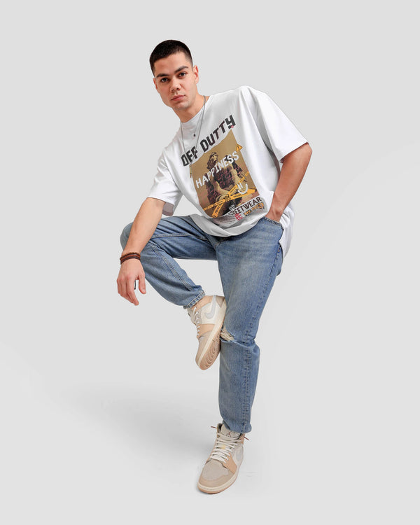 Front profile photo with a dynamic pose of a white Turkish look a like male model in a light grey backdrop wearing 'What Is Happiness' white relaxed T-shirt from Off Dutty's 'Rare and Radiant' collection, paired with striped jeans and an Air Jordan 1 Mid 'Milan' sneaker. He accessorizes with necklaces, three bracelets on his right hand.